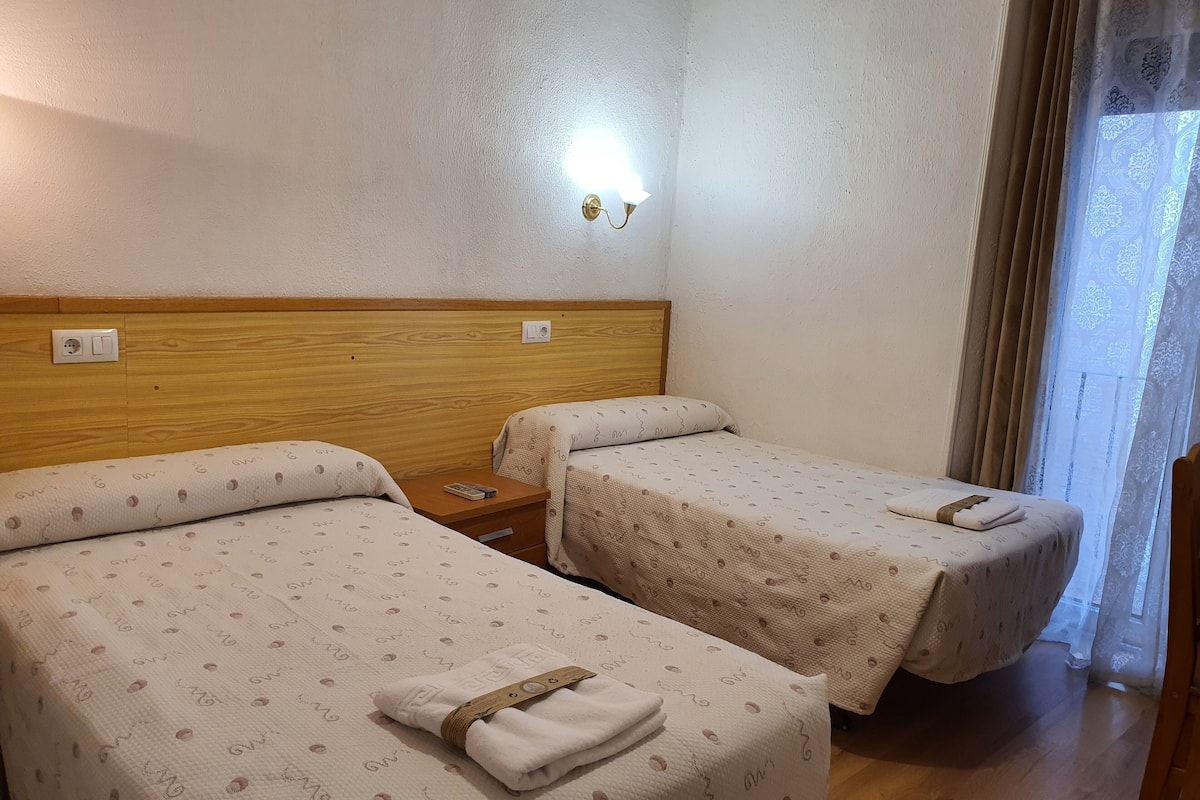 A hotel room with two single beds, wooden headboards, a nightstand with a remote control, and a wall-mounted light.