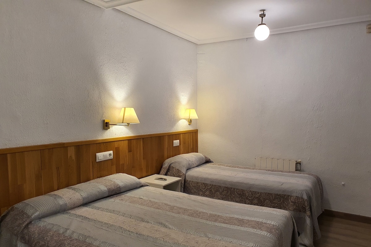 A room with two beds, wooden headboard, two wall-mounted lamps, a ceiling light, and a radiator.