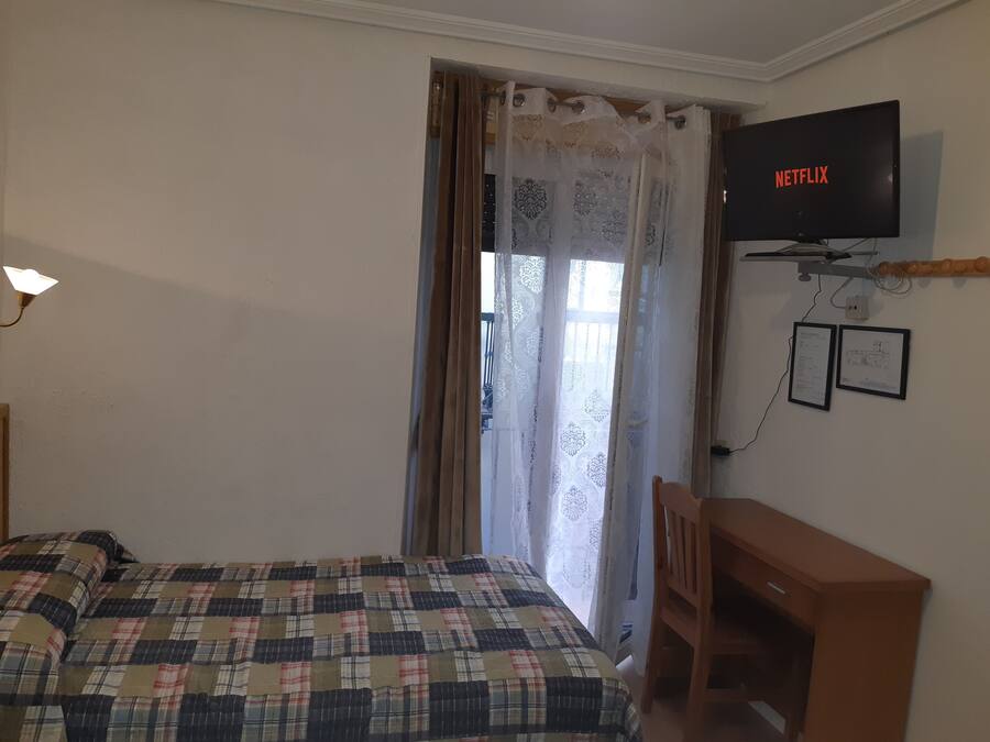 Un dormitorio con cama, televisor montado en la pared, escritorio y ventana con cortinas.