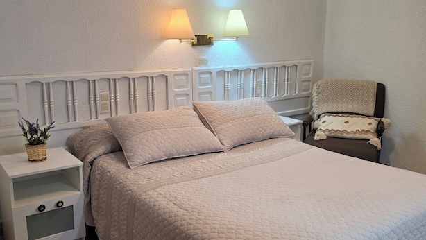 A bedroom with a white bed, a nightstand, a lamp, and a chair. - Pensión Martín (Jaén)