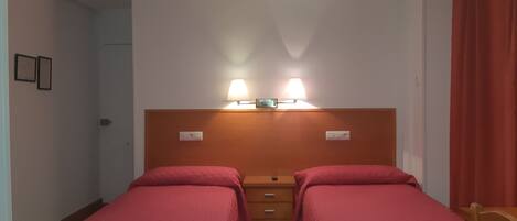 Habitación con dos camas individuales, cabecero de madera, dos lámparas de noche, una lámpara de techo y una puerta a la izquierda.