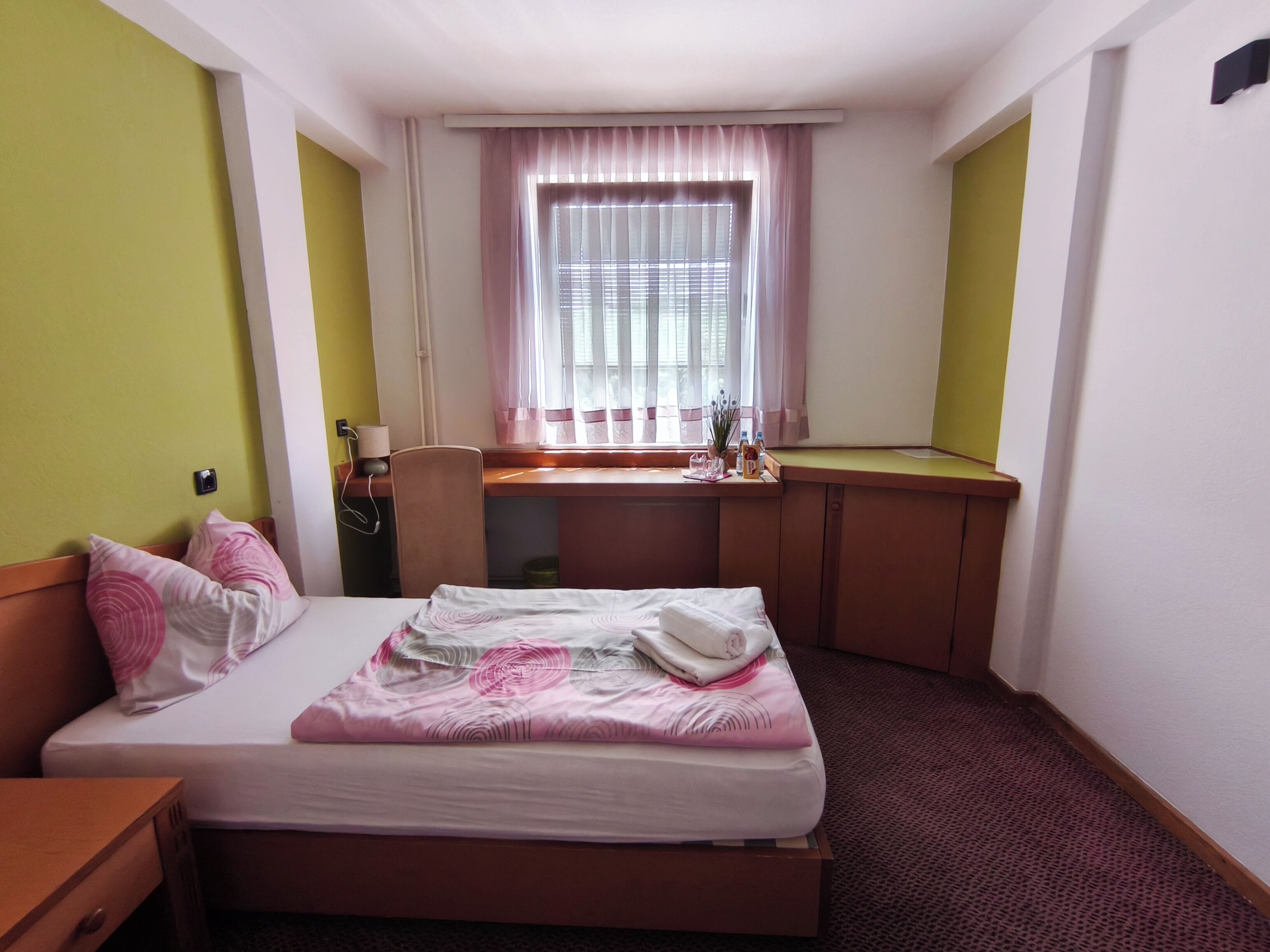Una camera d'hotel con un letto, una scrivania, una sedia, una finestra con tende e un comodino con una lampada.