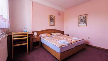 A hotel room with a single bed, a wooden nightstand, a chair, a desk, and two framed pictures on the walls.