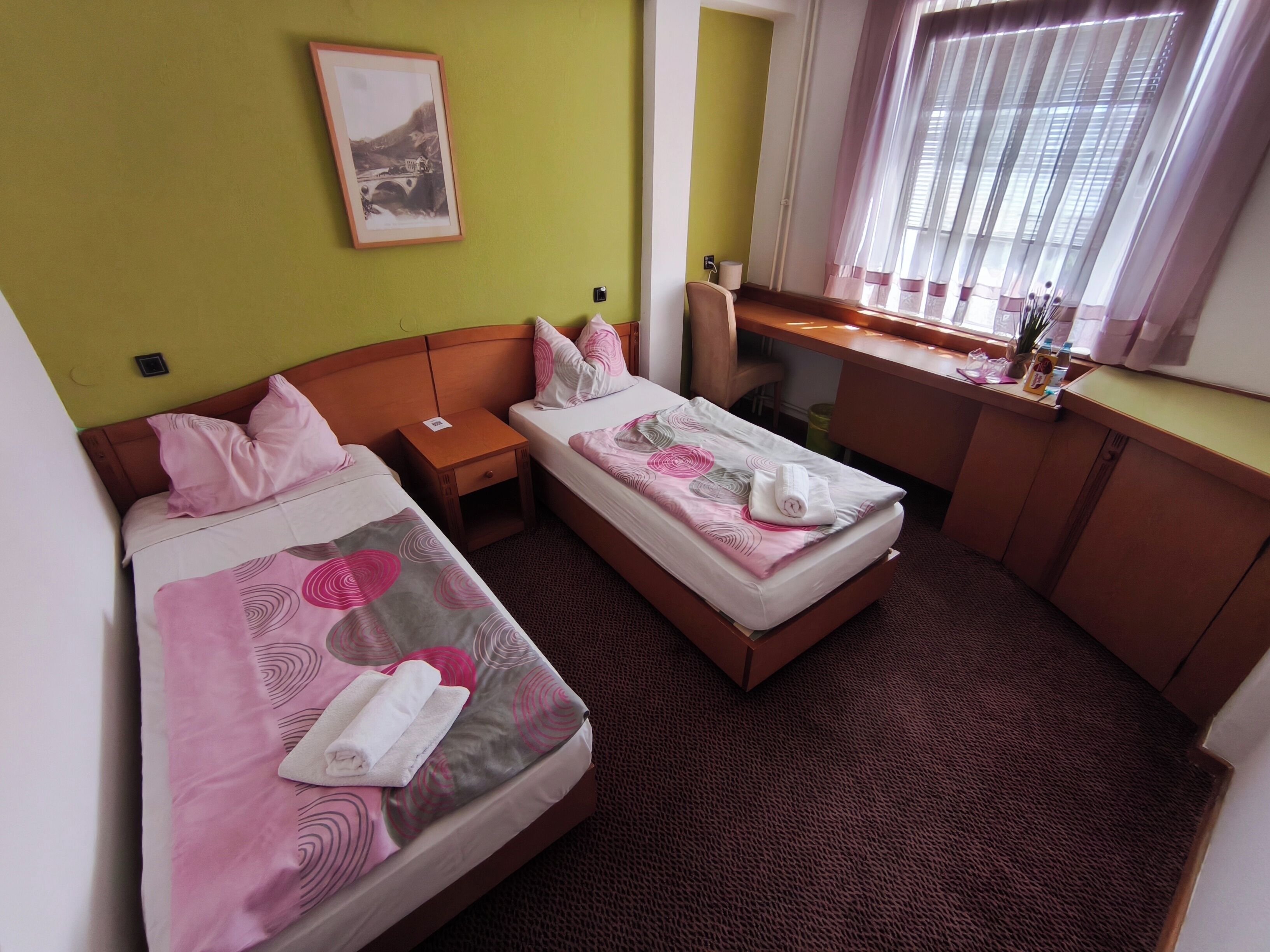 Ett hotellrum med två sängar, ett skrivbord, en stol, ett fönster med gardiner och en inramad tavla på väggen.