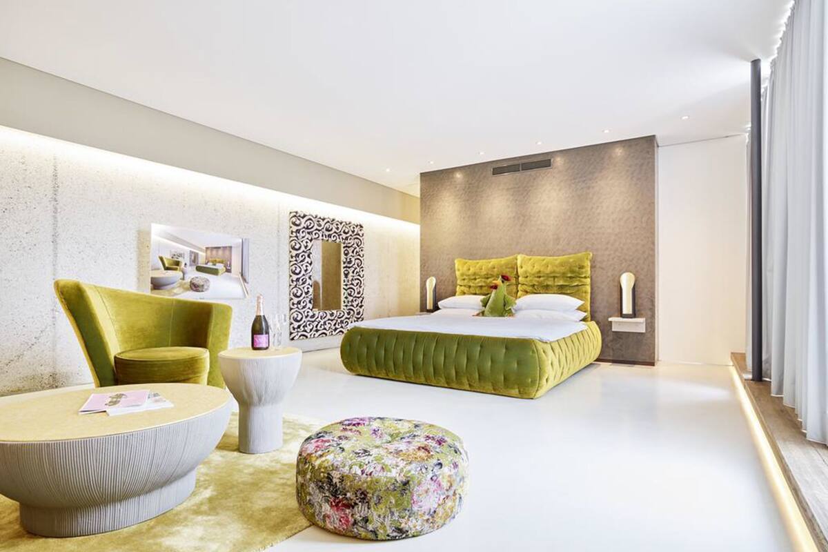 A modern bedroom with a green bed, a yellow armchair, a round ottoman, and a white round table.