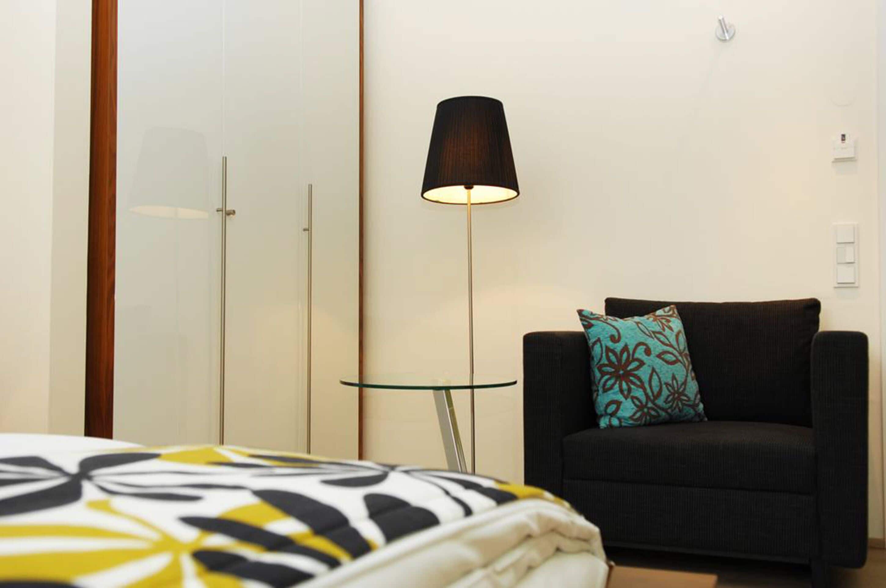 A hotel room with a bed, a black armchair with a patterned pillow, a floor lamp, and a small glass table.
