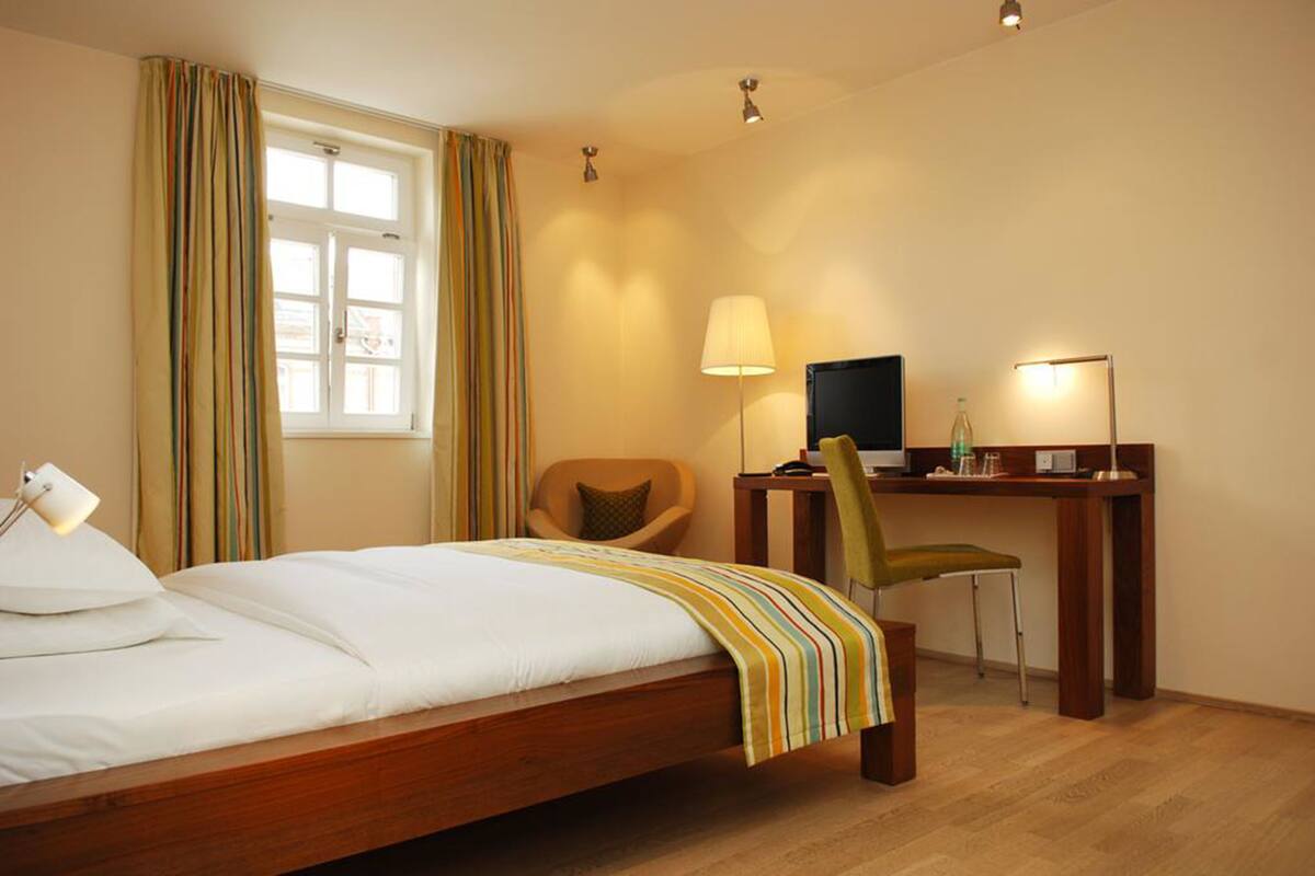 A hotel room with a bed, a desk with a computer, a chair, and a lamp.