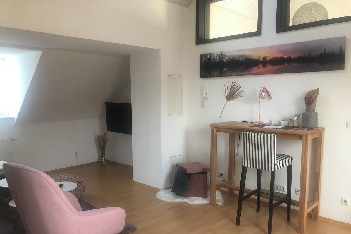 A modern living room with a pink chair, a wooden desk with two striped chairs, a flat-screen TV, and artwork on the wall.