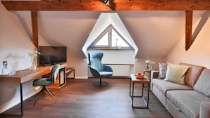 A modern loft bedroom with a sloped ceiling, wooden beams, a large window, a TV, a desk, a chair, a sofa, and a small table. - Hotel-Gasthof zum Bach (Neukirchen Beim Heiligen Blut)