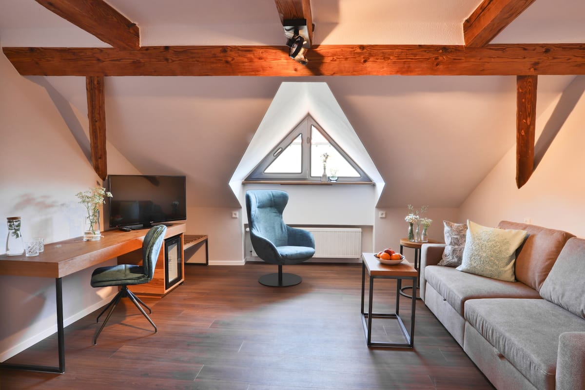 A modern loft bedroom with a sloped ceiling, wooden beams, a large window, a TV, a desk, a chair, a sofa, and a small table.