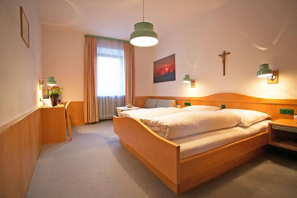 A hotel room with a wooden bed, a desk with a lamp, a chair, a window with curtains, and wall-mounted lights.