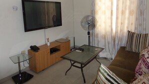 A living room with a flat-screen TV, a standing fan, a glass-top coffee table, a wooden cabinet, and a brown sofa with striped cushions. - House No 12B (Lagos)