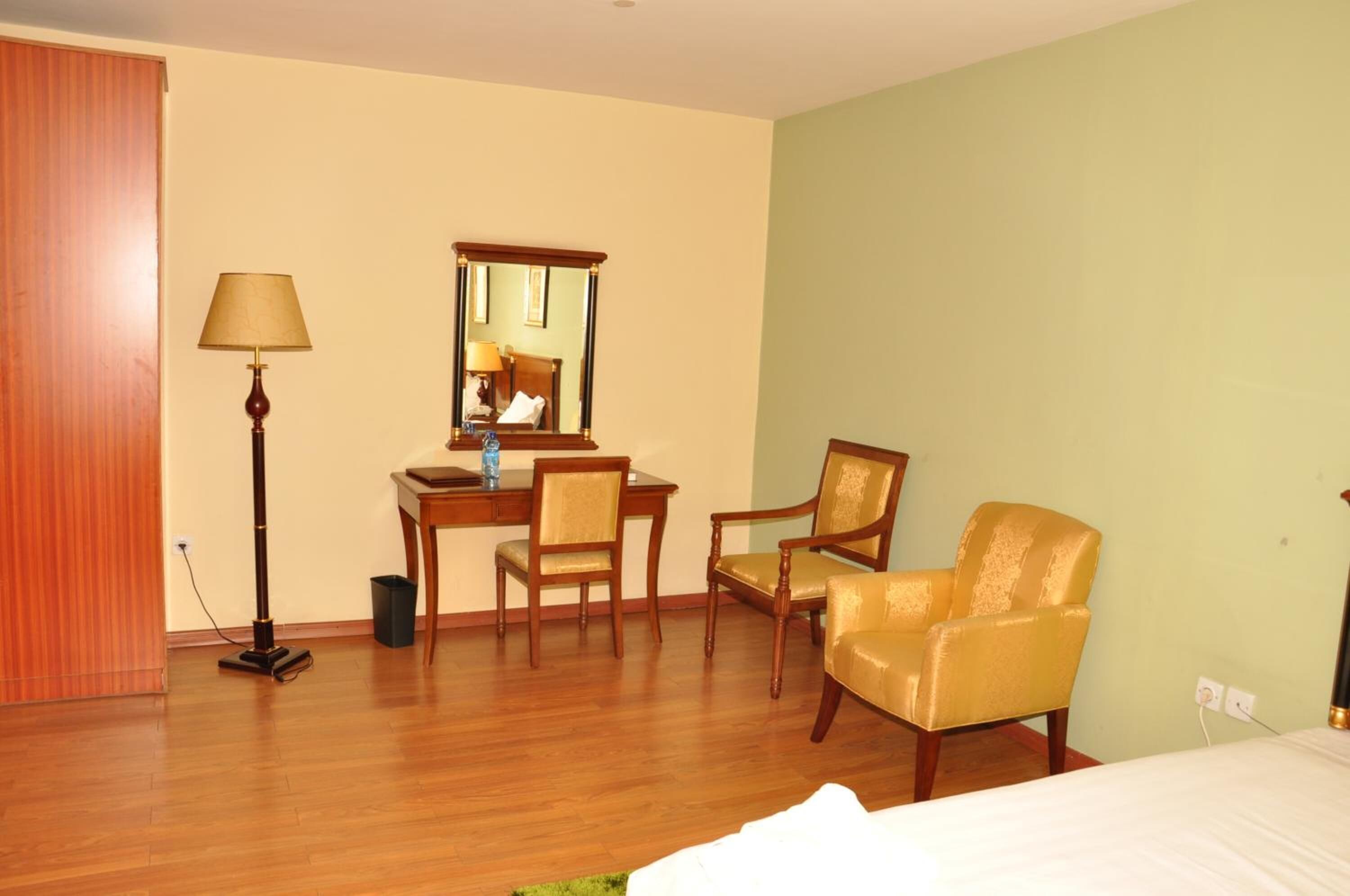 A hotel room with a wooden floor, a desk with a chair, a bed, a lamp, and a mirror.
