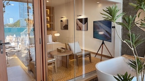 Habitació d'hotel amb una porta de vidre que dona a un balcó amb vista al mar i edificis. A l'interior, hi ha un llit, un escriptori, una cadira, una làmpada i un quadre sobre un trípode.