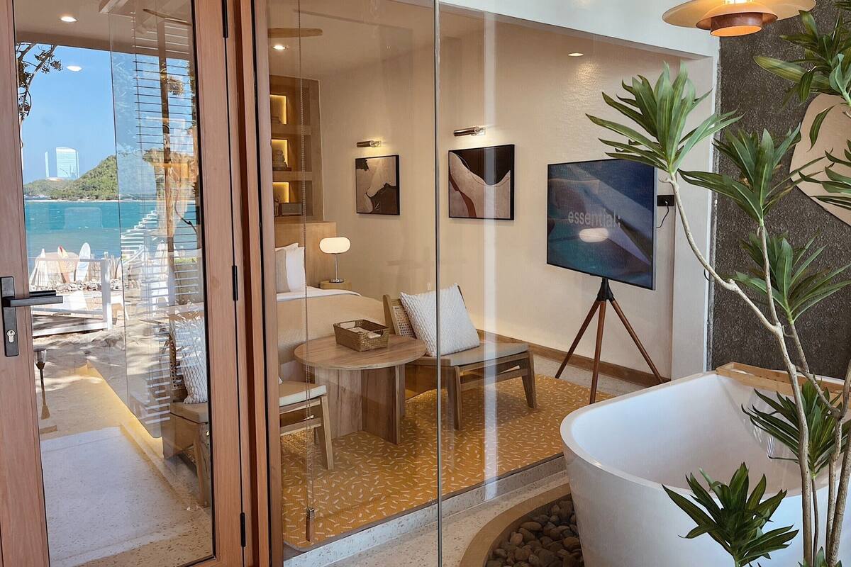 Habitación de hotel con puerta de vidrio que da a un balcón con vista al mar y edificios. En el interior, hay una cama, un escritorio, una silla, una lámpara y un cuadro sobre un atril.