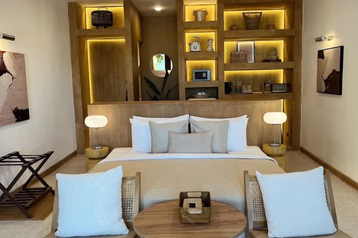 A hotel room with a large bed, two bedside tables with lamps, a small round table with a basket, and shelves with decorative items.