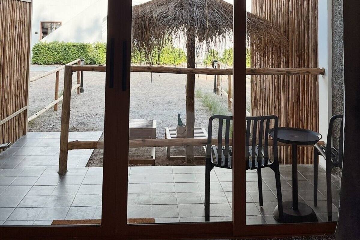 Una habitación con un gran ventanal corredero que da a un espacio exterior con un paraguas de paja, vallas de madera y vistas a un edificio en la distancia.