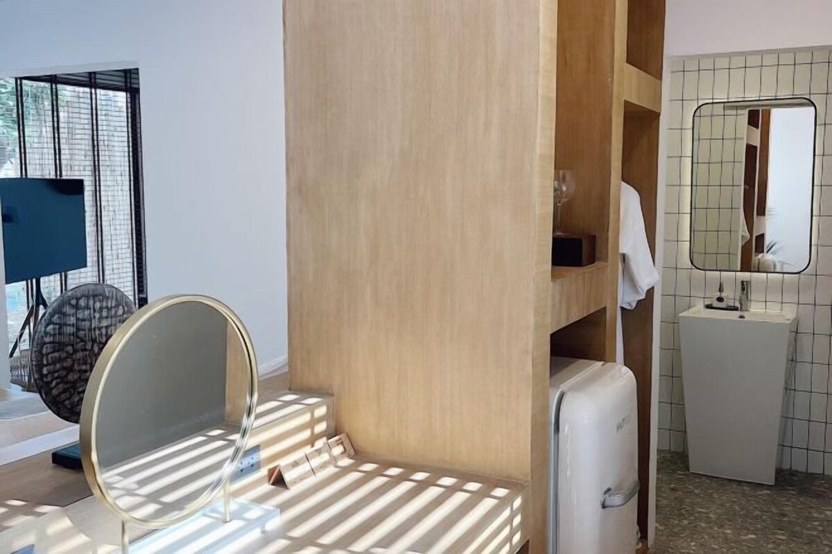 A compact hotel room with a wooden vanity, a round mirror, a small sink, and a compact refrigerator.