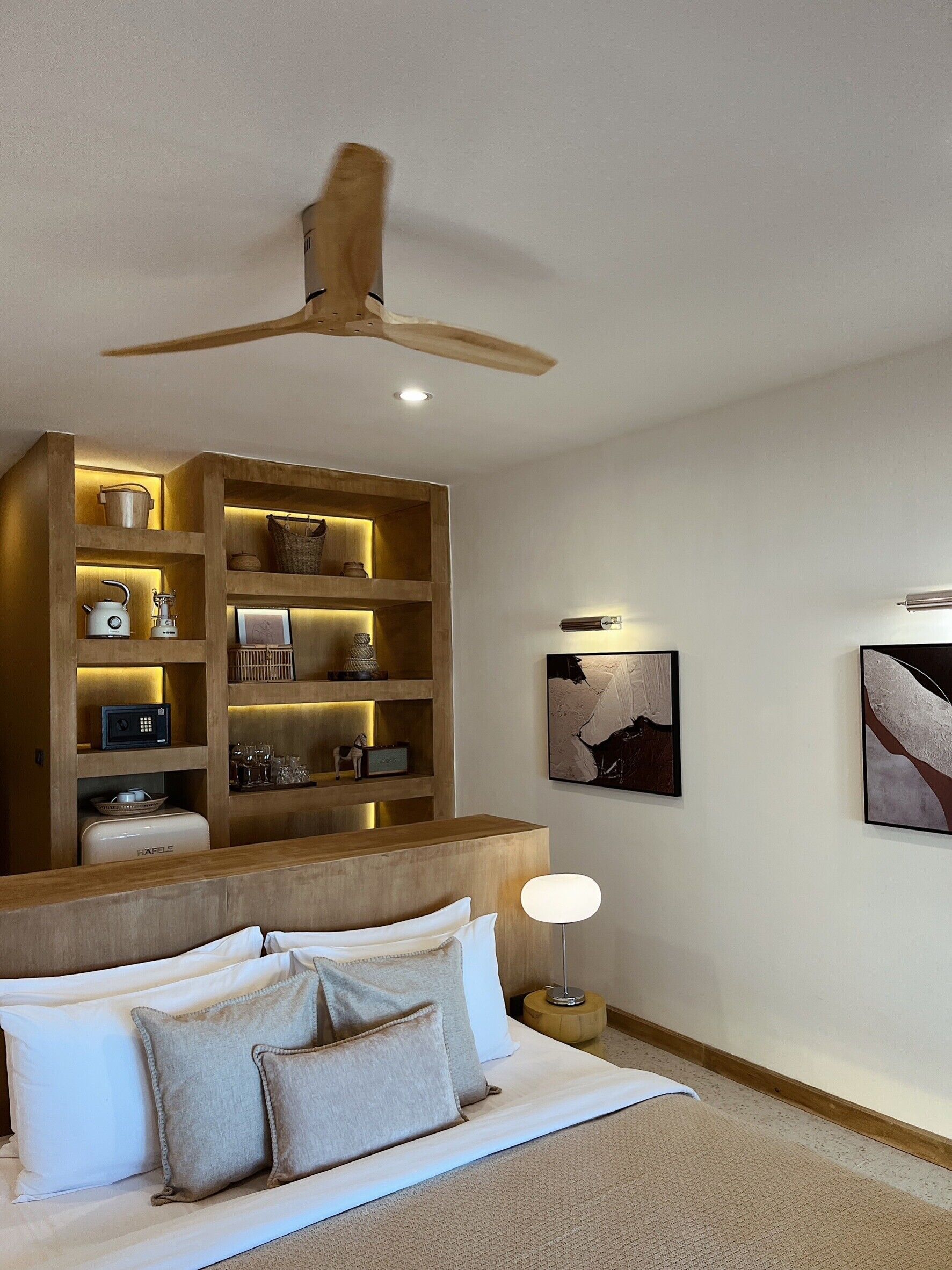 Une chambre à coucher moderne avec une tête de lit en bois, un ventilateur de plafond, une étagère intégrée contenant divers objets, un lit avec du linge de lit blanc et deux œuvres d’art accrochées au mur.