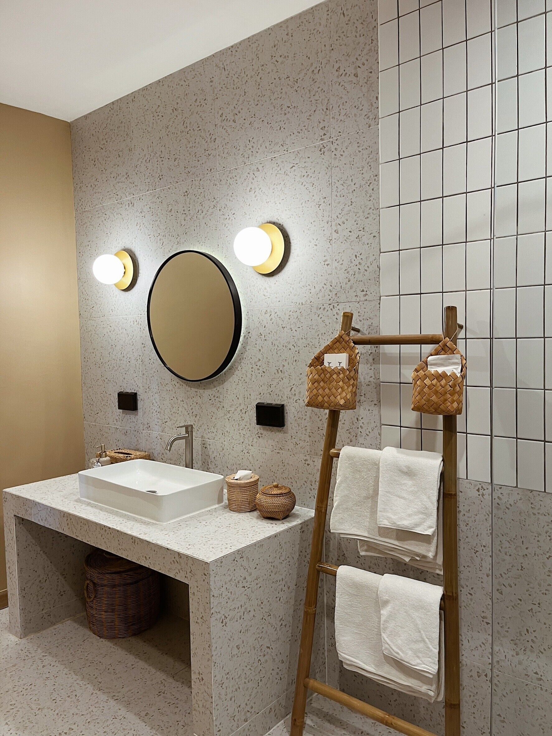 A bathroom with a freestanding sink, two wall-mounted lights, a round mirror, and a towel rack with multiple towels.