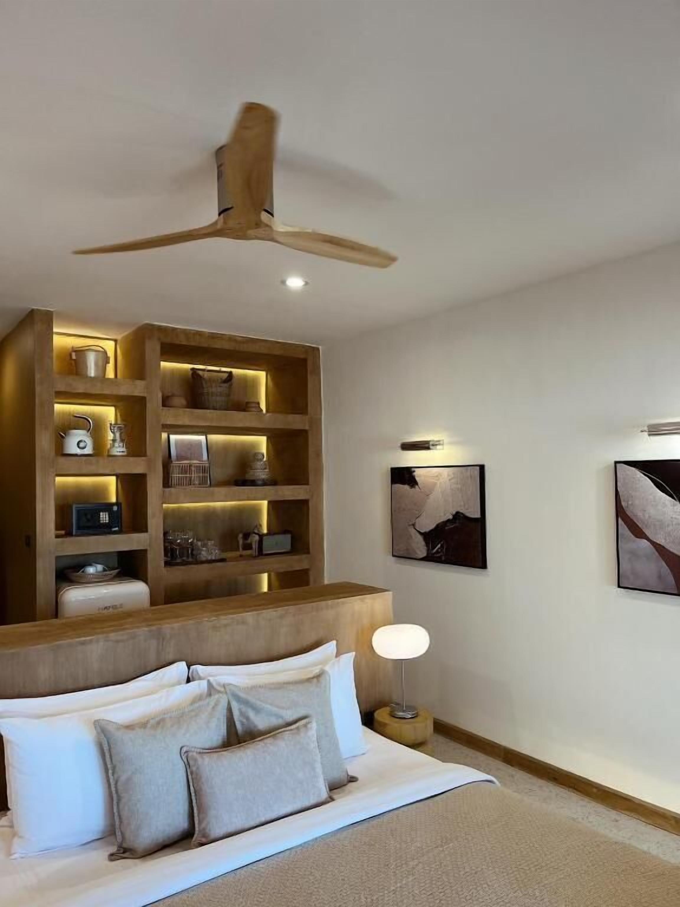A modern hotel room with a large bed, a wooden headboard, a ceiling fan, a built-in shelf unit with various items, and two abstract paintings on the wall.