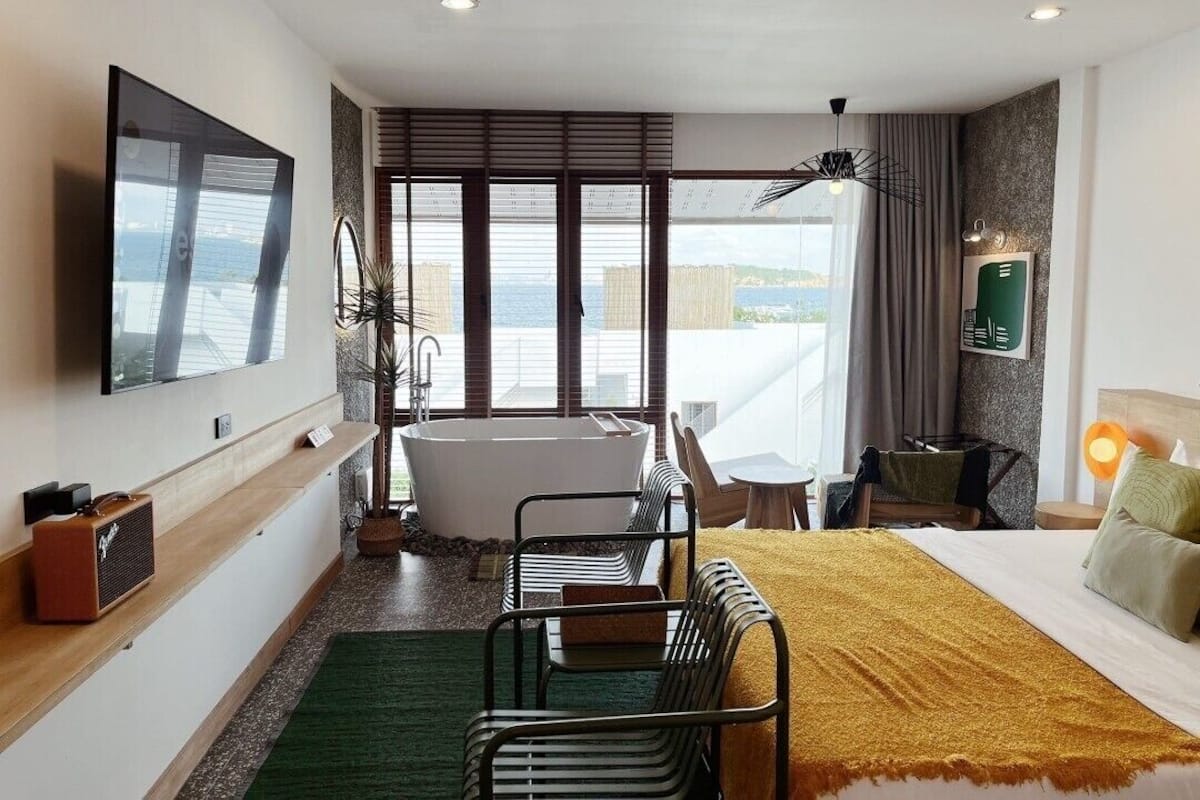 Habitación de hotel moderna con una cama grande, bañera, zona de estar con sillas, televisión y vista al mar.