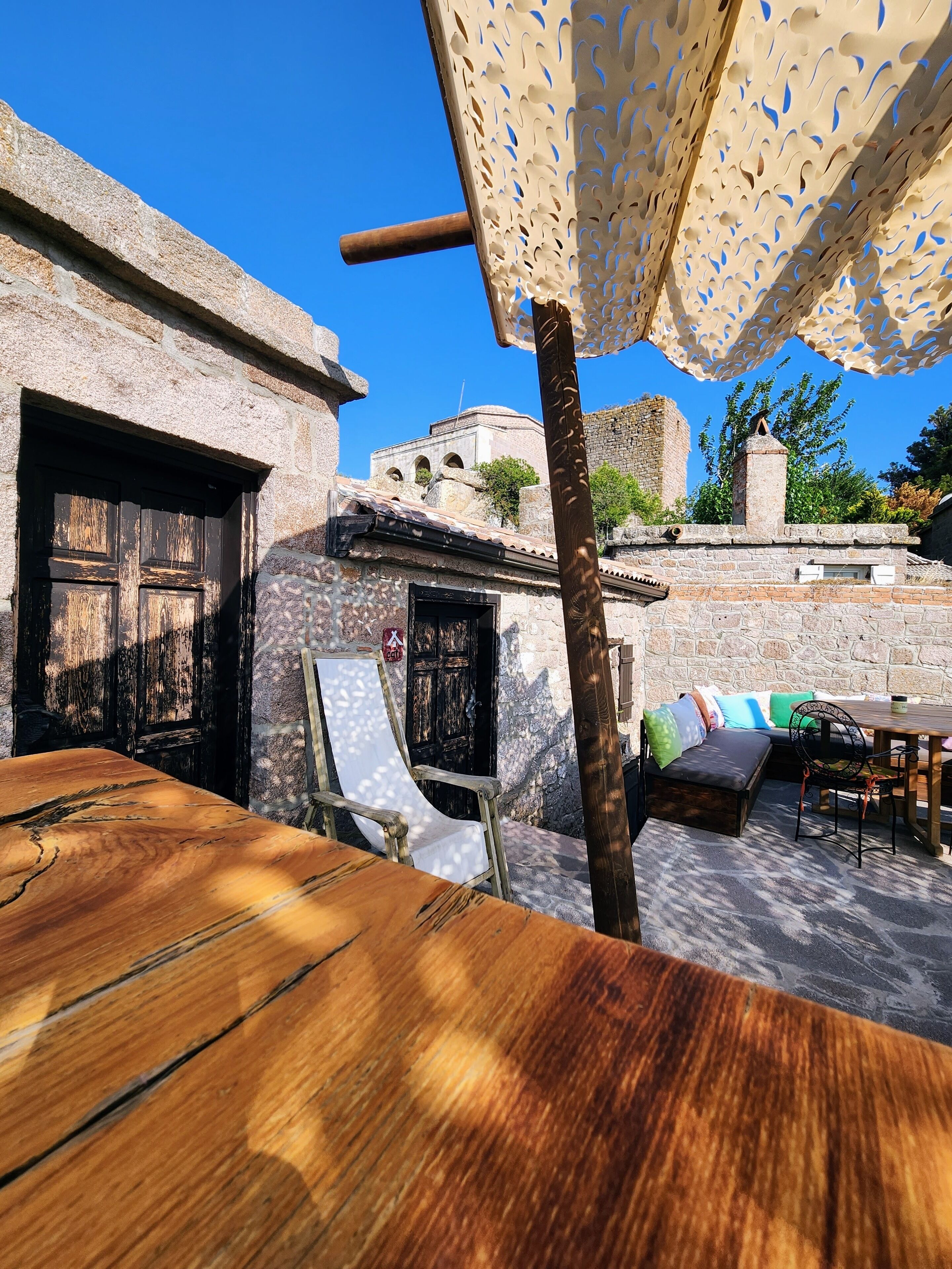 A rustic outdoor seating area with a wooden table and chairs, a patterned umbrella, and stone buildings in the background.