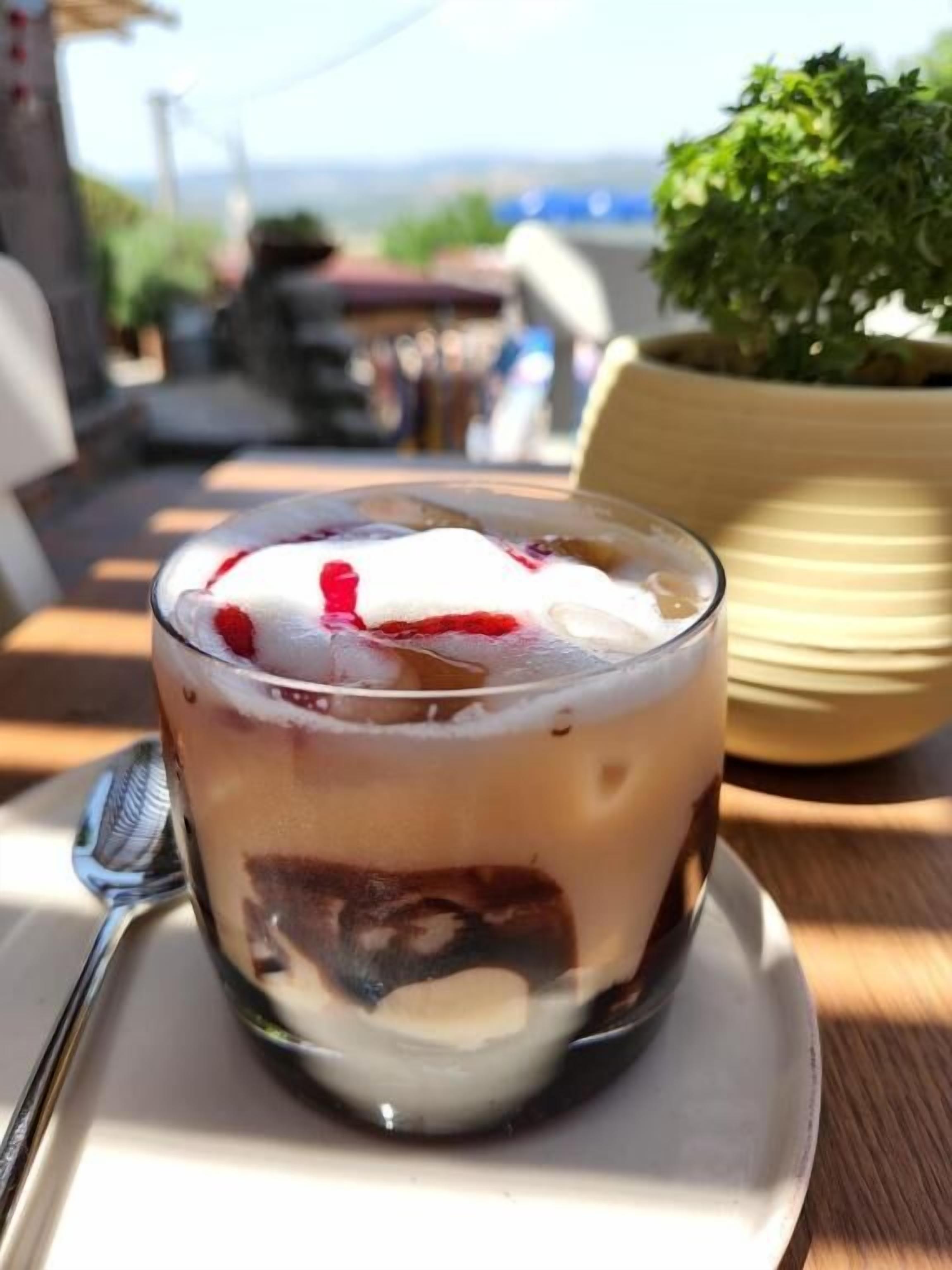 Postre en vaso con capas, servido en un plato blanco con una cuchara, sobre una mesa de madera con una planta en maceta al fondo.