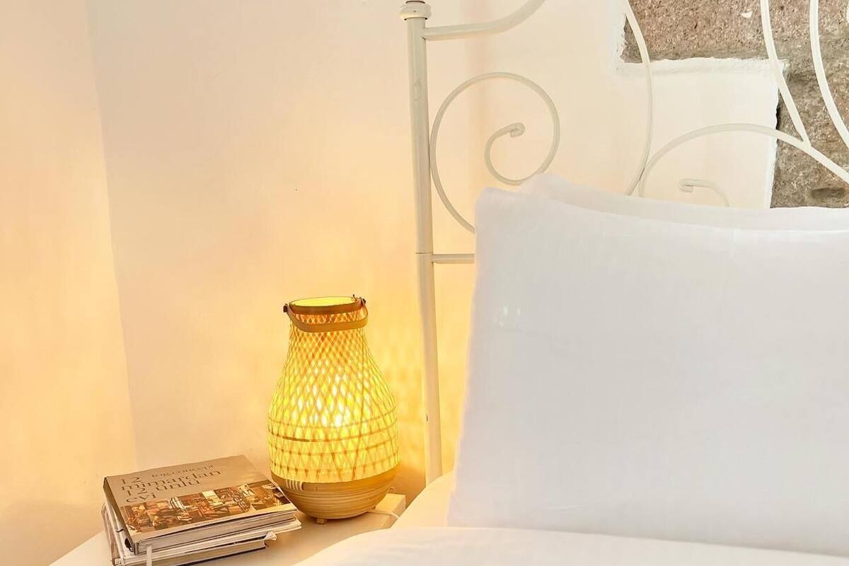 A neatly made bed with a white pillow and a patterned blanket, a small table with a lamp and a book, and a metal headboard.