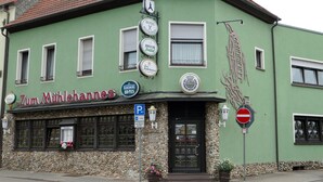 A green building with a sign for Zum Mühlehaus, featuring a clock, a sign for Bruch Zwickel, and a sign for Karlsberg URPILS. - Zum Mühlehannes (Sankt Ingbert)