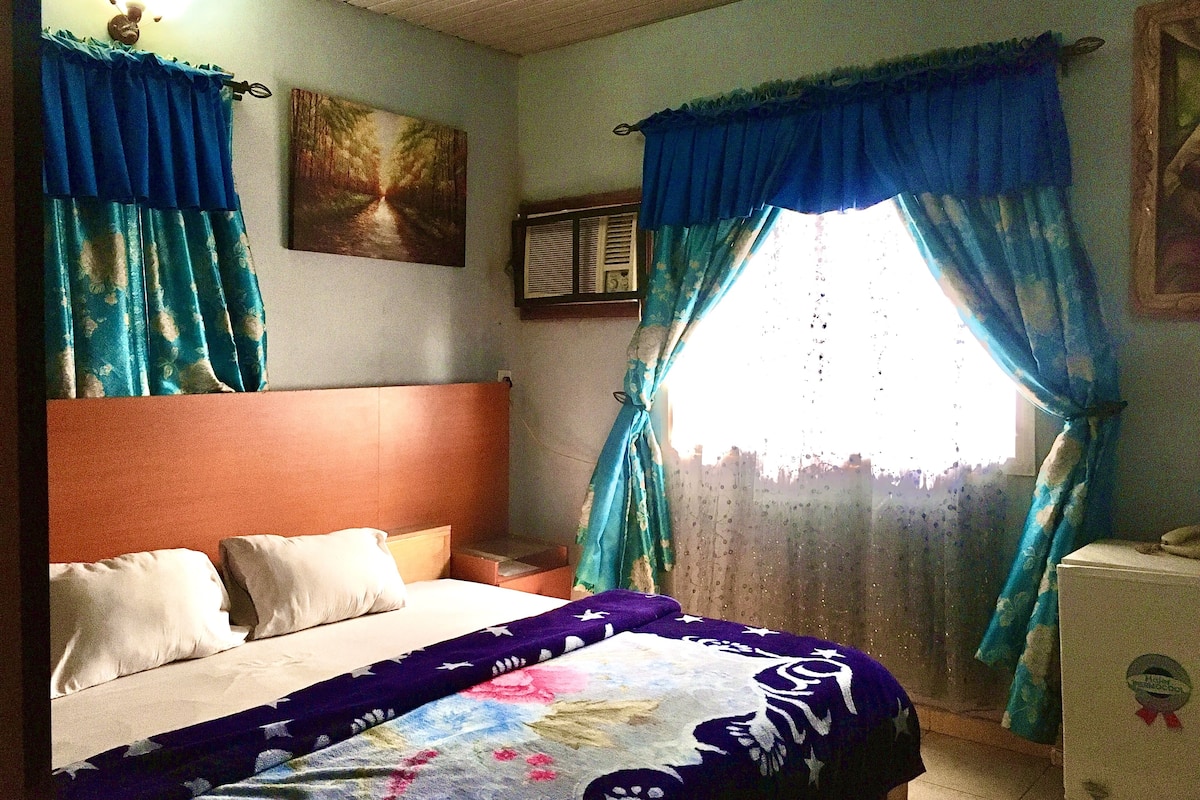 A bedroom with a bed, a headboard, curtains, a window, and a wall-mounted TV.