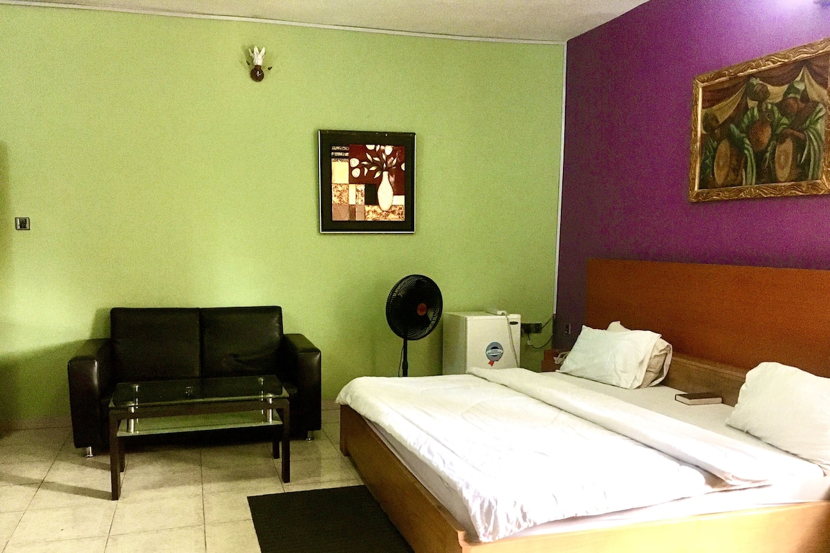 A hotel room with a bed, a sofa, a fan, and a painting on the wall.