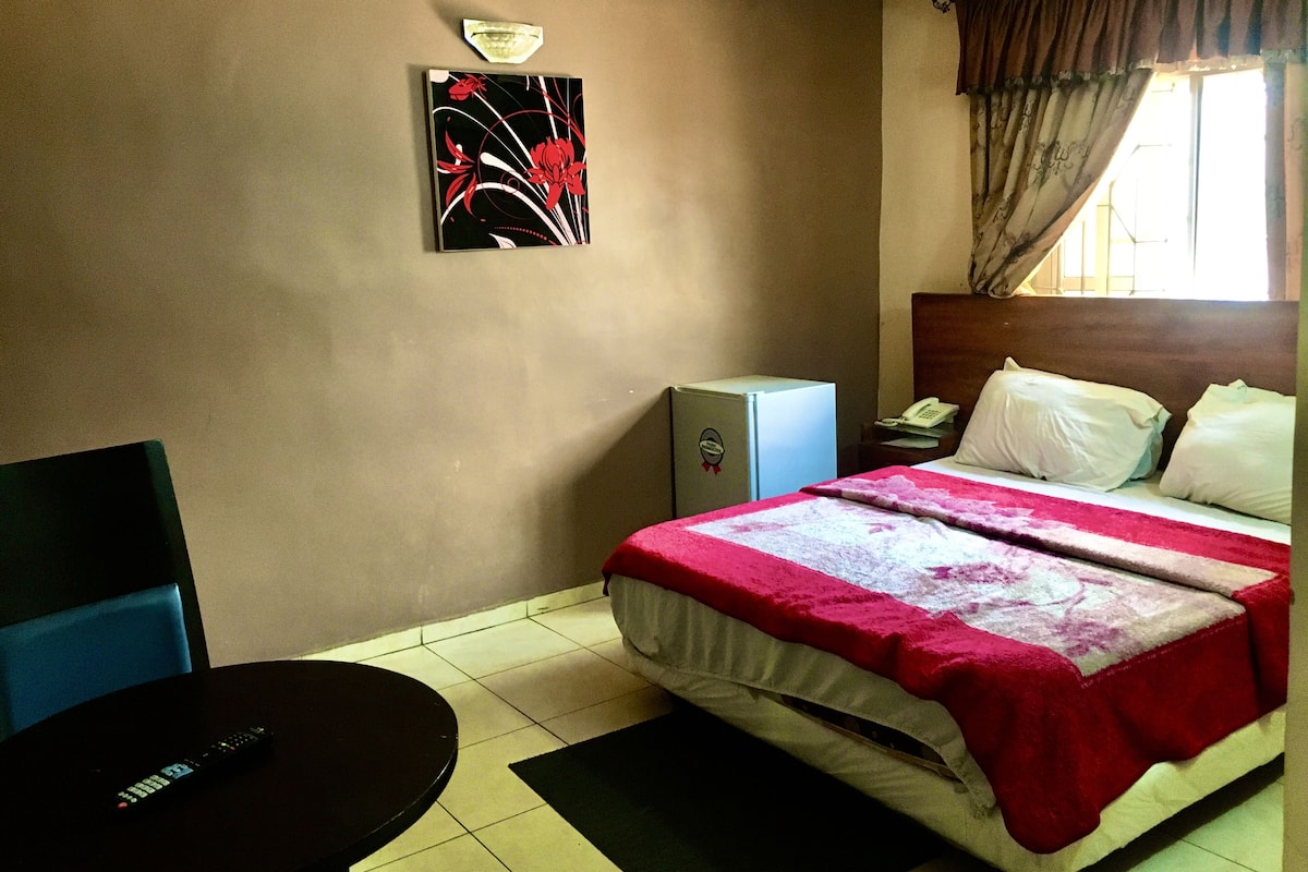 A hotel room with a bed, a desk, a chair, a refrigerator, and a wall art piece.