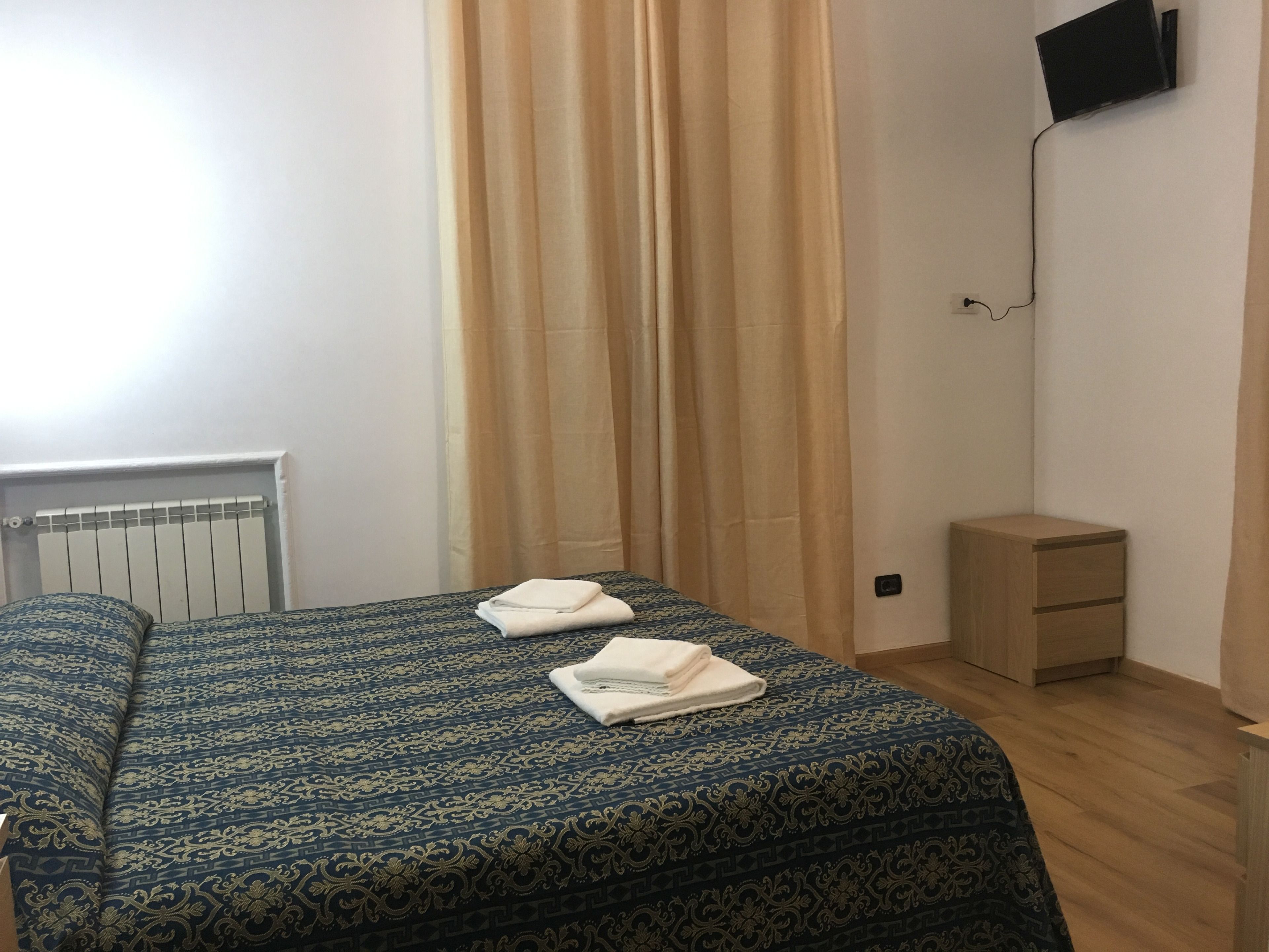 Una habitación con cama, mesita de noche, televisor montado en la pared y radiador.