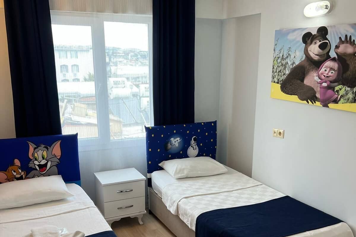 Une chambre d’hôtel avec deux lits, une table de chevet, une fenêtre avec des rideaux et un mur orné d’une affiche représentant un personnage de dessin animé.