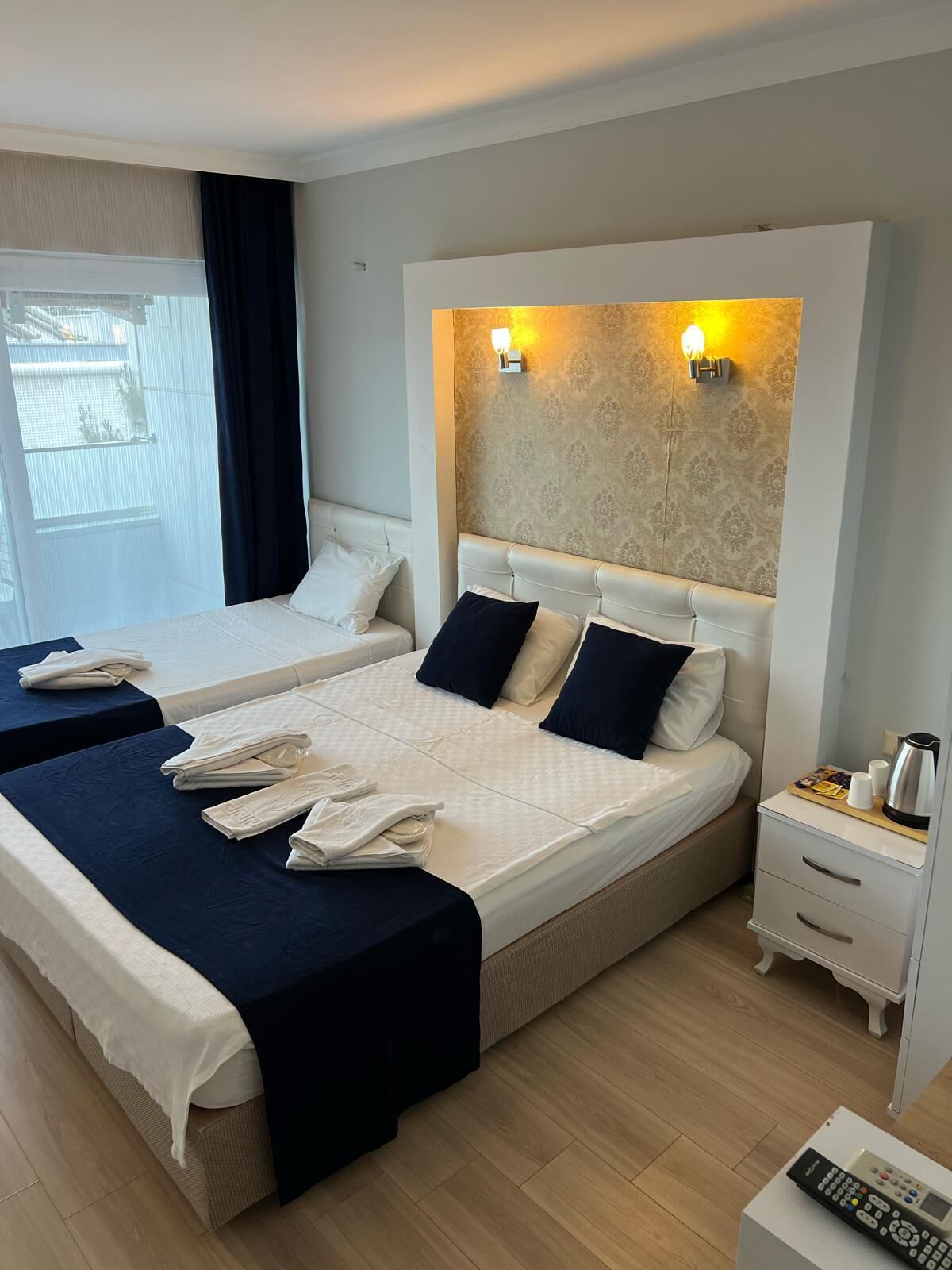A hotel room with two beds, a nightstand with a remote control, and a window with curtains.