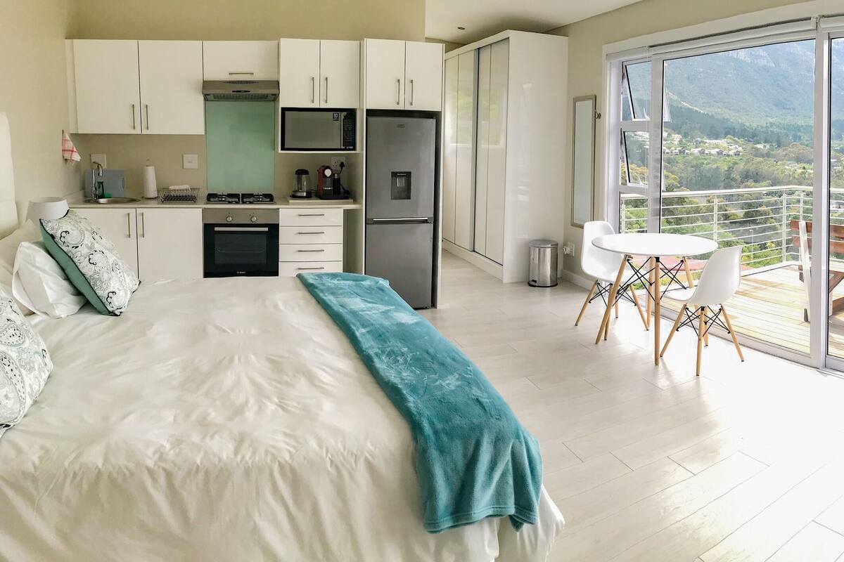 Un dormitorio con una cama grande, un balcón con mesa y sillas, y una zona de cocina.