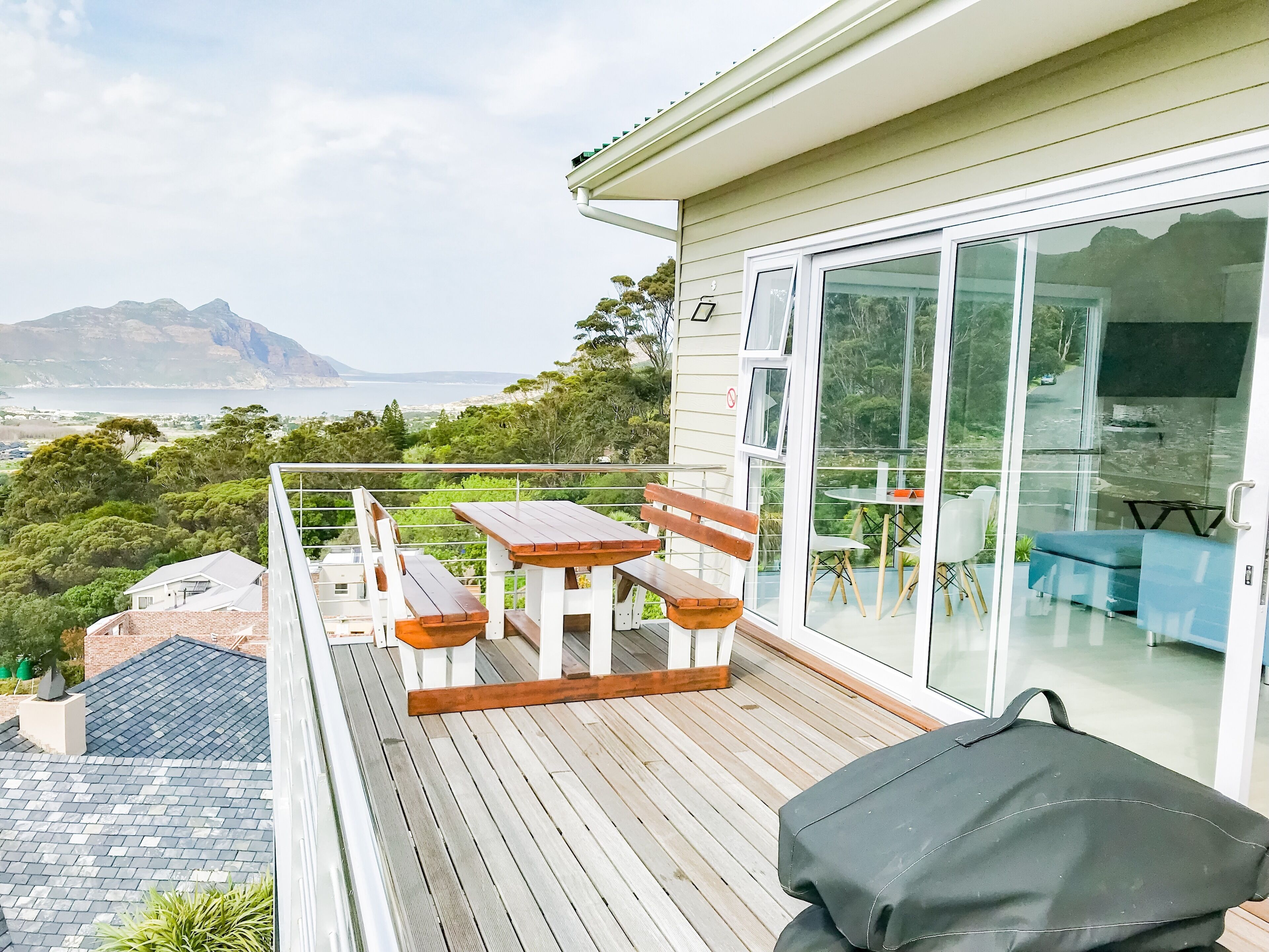 A balcony with wooden furniture, a grill, and a view of the sea and mountains.