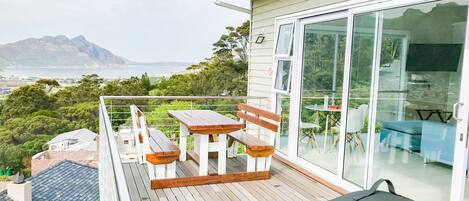 A balcony with wooden furniture, a grill, and a view of the sea and mountains.