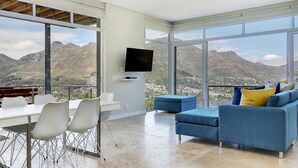 A modern living room with a large window, a flat-screen TV, a dining area with chairs, and a blue sofa with cushions. - Mount Bay (Cape Town)
