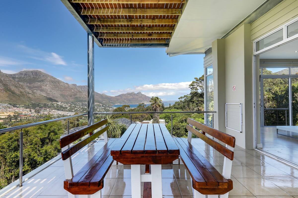 Mesa y bancos de madera en un balcón con vistas a montañas y una ciudad.