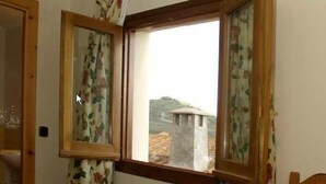 A room with a wooden-framed window, floral curtains, and a view of a stone structure outside. - Hostal Las Grullas (Tornos)