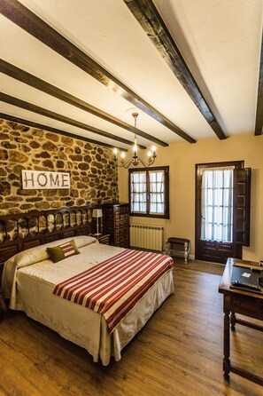 Un dormitorio con pared de piedra, cama de madera con colcha a rayas rojas y blancas, un candelabro, mesita de noche de madera y una ventana con cortinas.