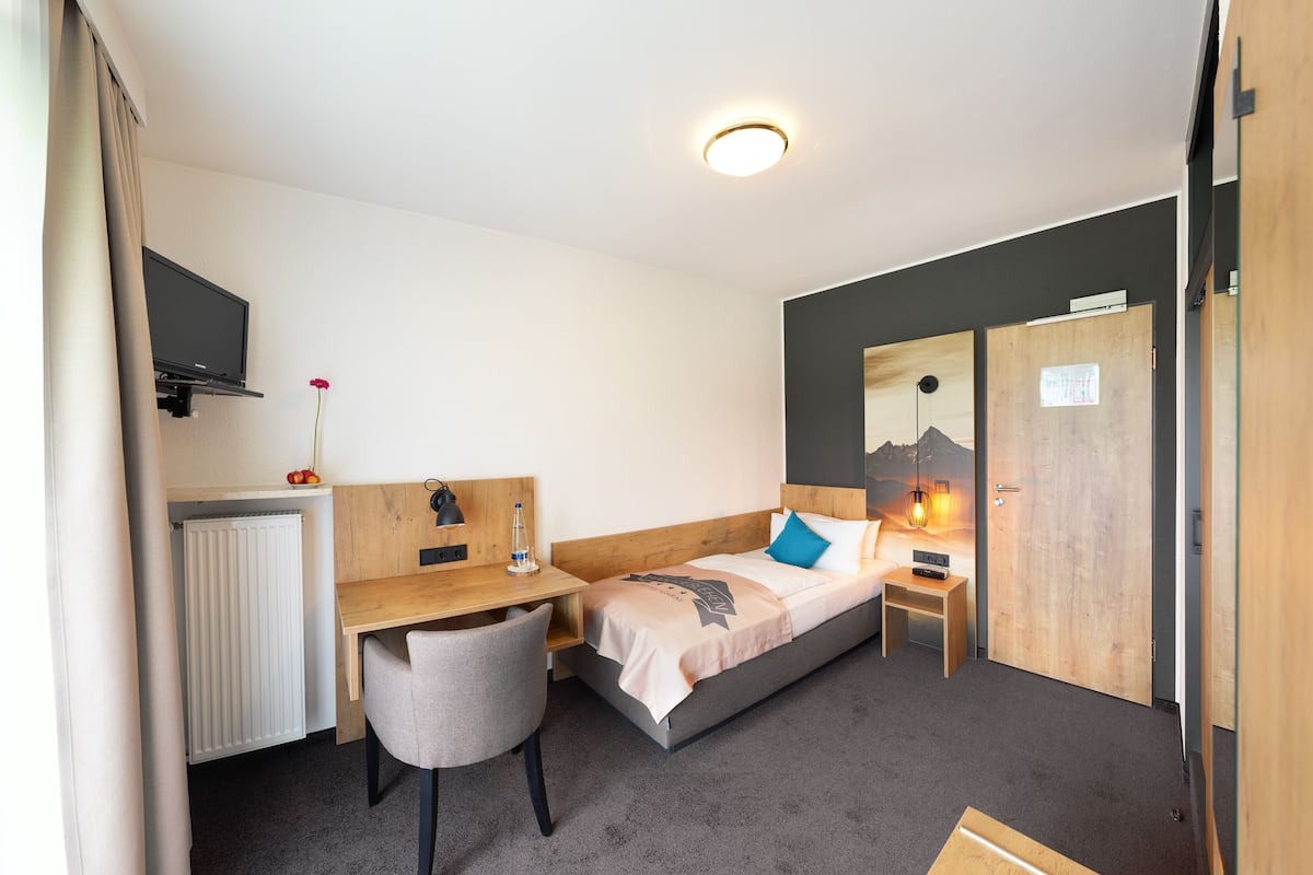 A hotel room with a bed, a desk, a chair, a radiator, a TV, and a door.