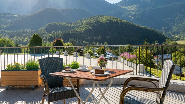 A terrace with a table and chairs, overlooking a mountainous landscape. - Hotel Garni Hanauerlehen (Schoenau am Koenigssee)