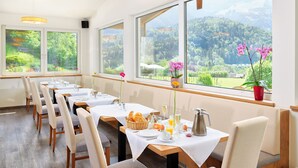 A dining area with tables set for breakfast, large windows with a mountain view, and a wooden ceiling. - Hotel Garni Hanauerlehen (Schoenau am Koenigssee)
