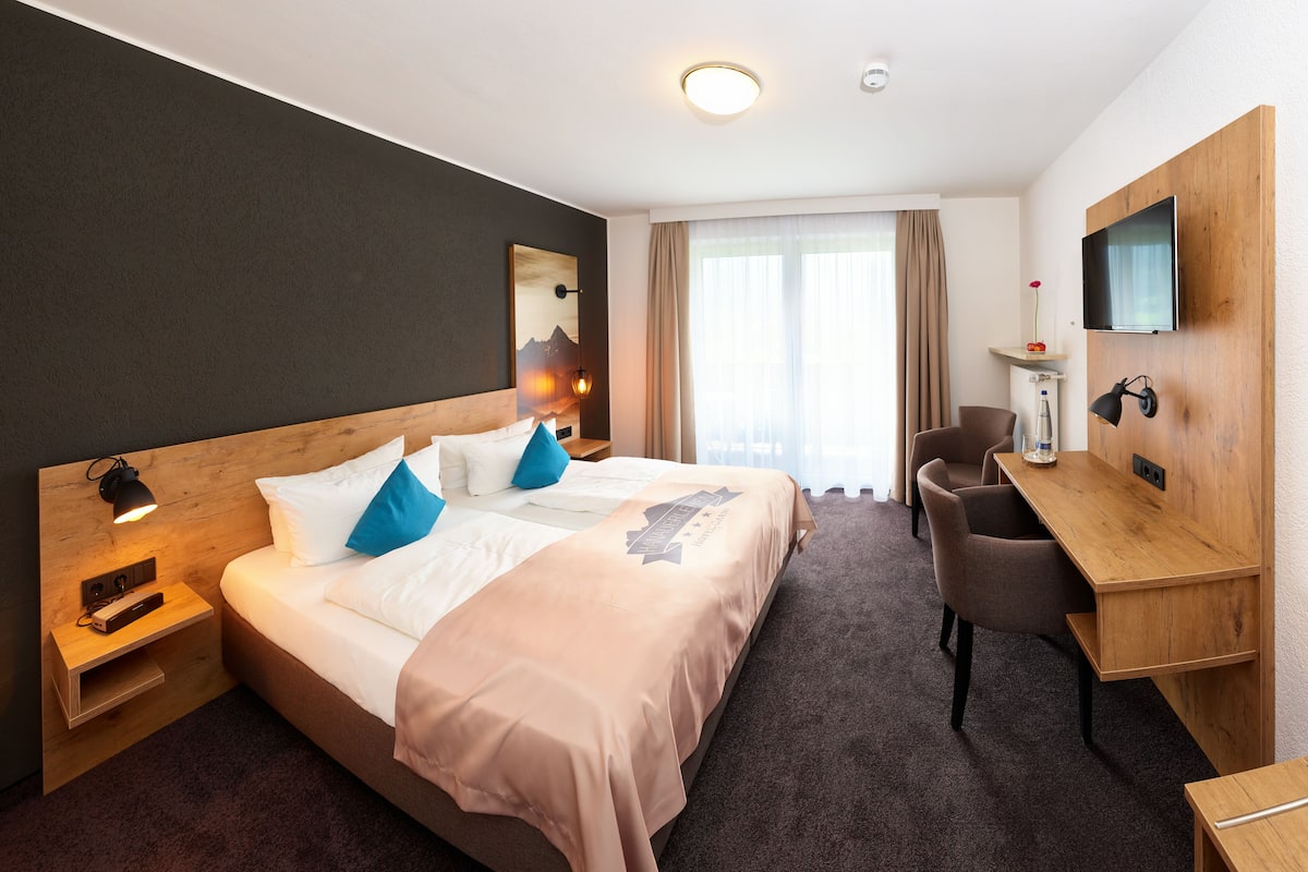 A hotel room with a large bed, a desk, a chair, a TV, and a window with curtains.