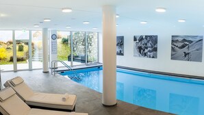 A modern indoor swimming pool with clear glass walls, a pool ladder, and lounge chairs with towels. - Hotel Garni Hanauerlehen (Schoenau am Koenigssee)