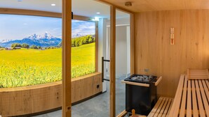 A wooden sauna with a bench and a stove, a wooden wall, and a large window with a view of a green field and mountains. - Hotel Garni Hanauerlehen (Schoenau am Koenigssee)