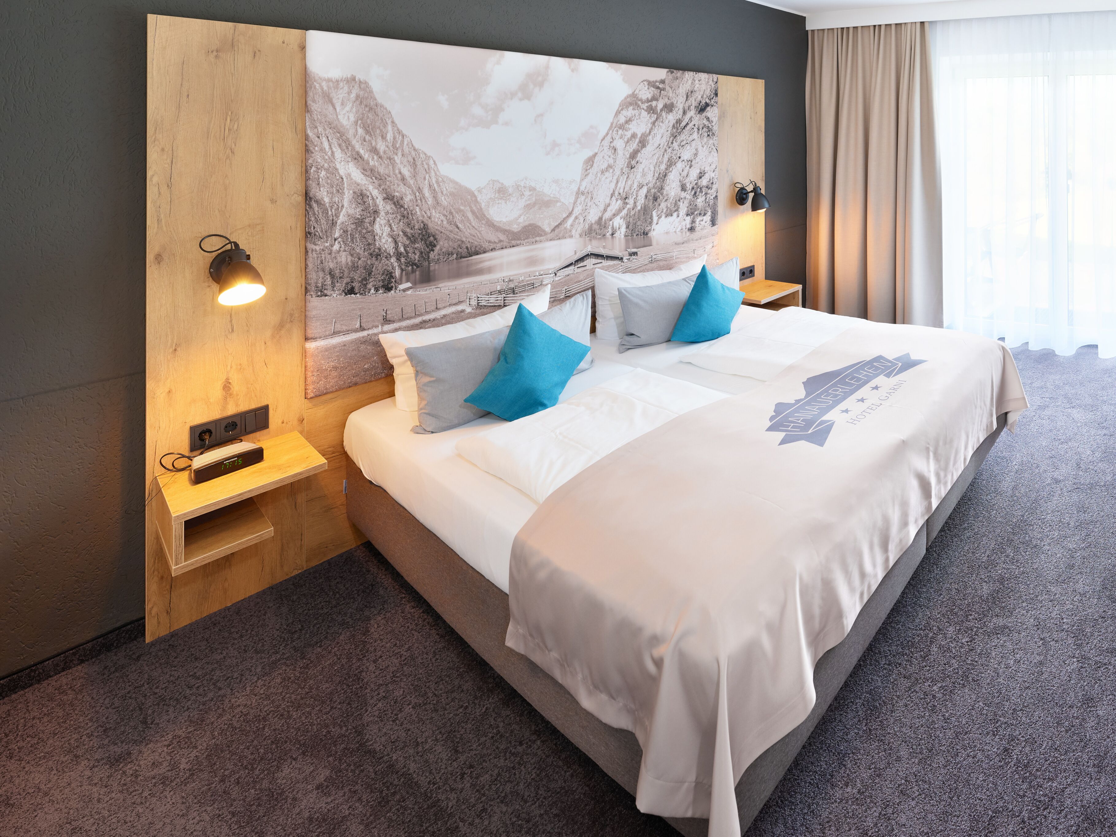 A hotel room with a large bed, a headboard with a scenic print, a bedside table with a lamp, and a nightstand with a clock.